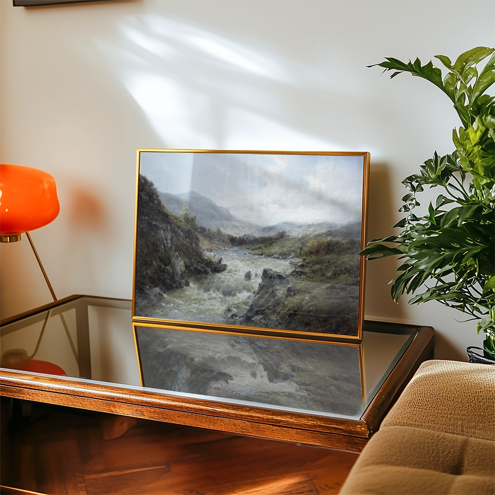 A serene landscape painting of a river flowing through rolling hills, framed elegantly and displayed on a coffee table next to a contemporary lamp and a plant.
