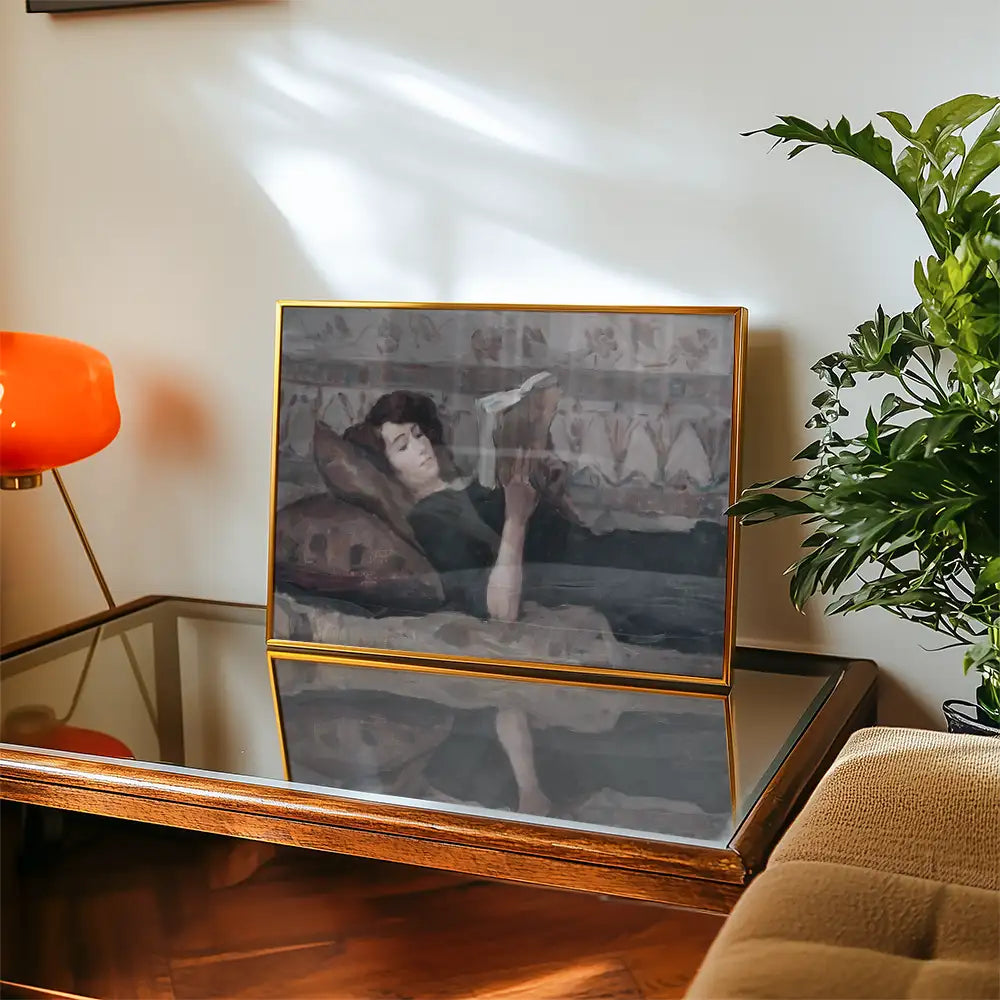Painting of a woman reading while lying on a couch, displayed in a gold frame on a wooden table with a lamp and plant in the background.