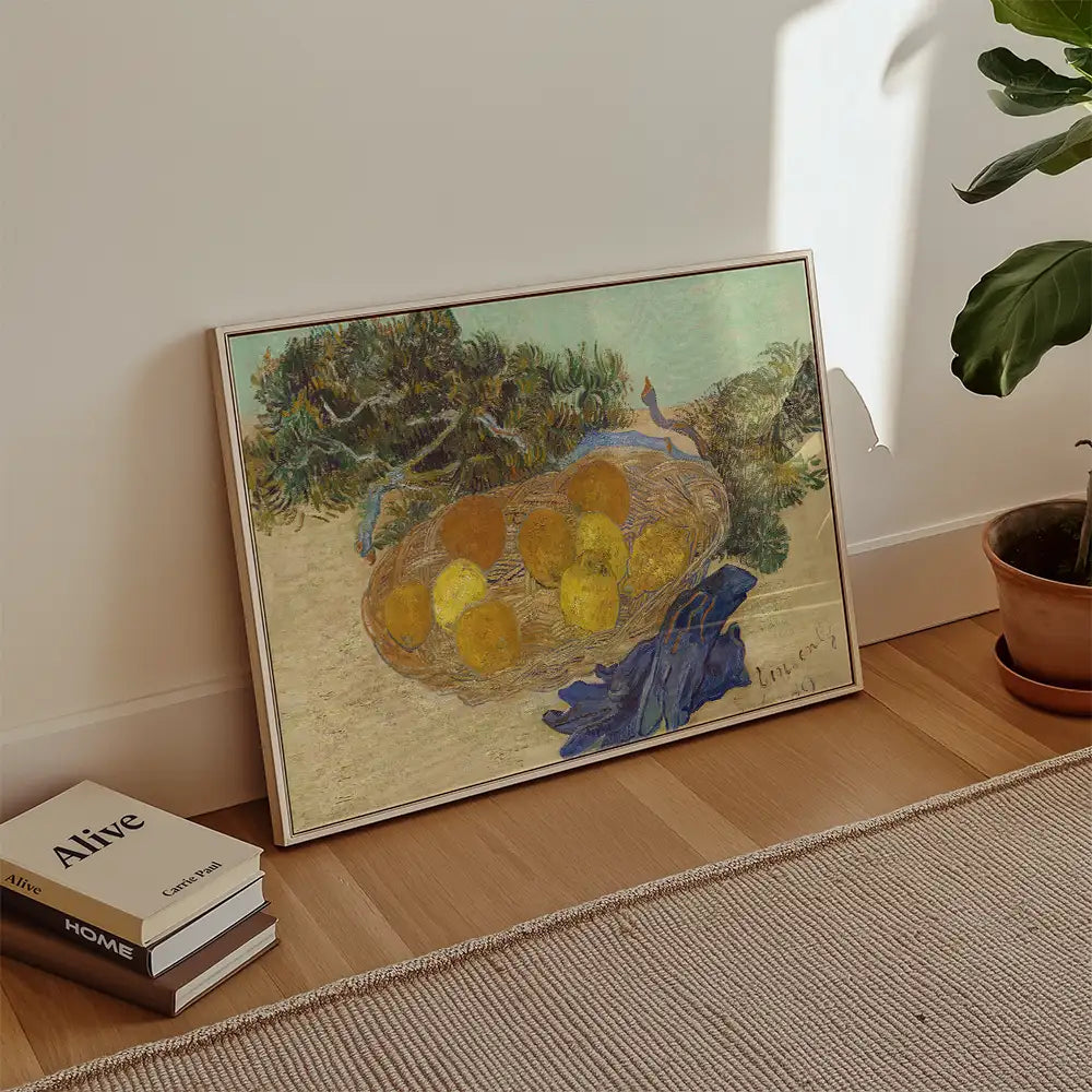 Framed painting of a basket with lemons and a blue cloth, placed on a wooden floor next to books and a potted plant.