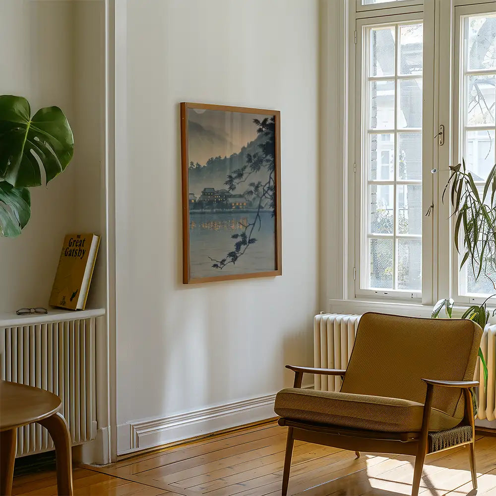 A serene landscape painting framed on a wall in a cozy living room, featuring a view of water and trees, accompanied by a brown armchair and a bookshelf with the book "Great Gatsby."