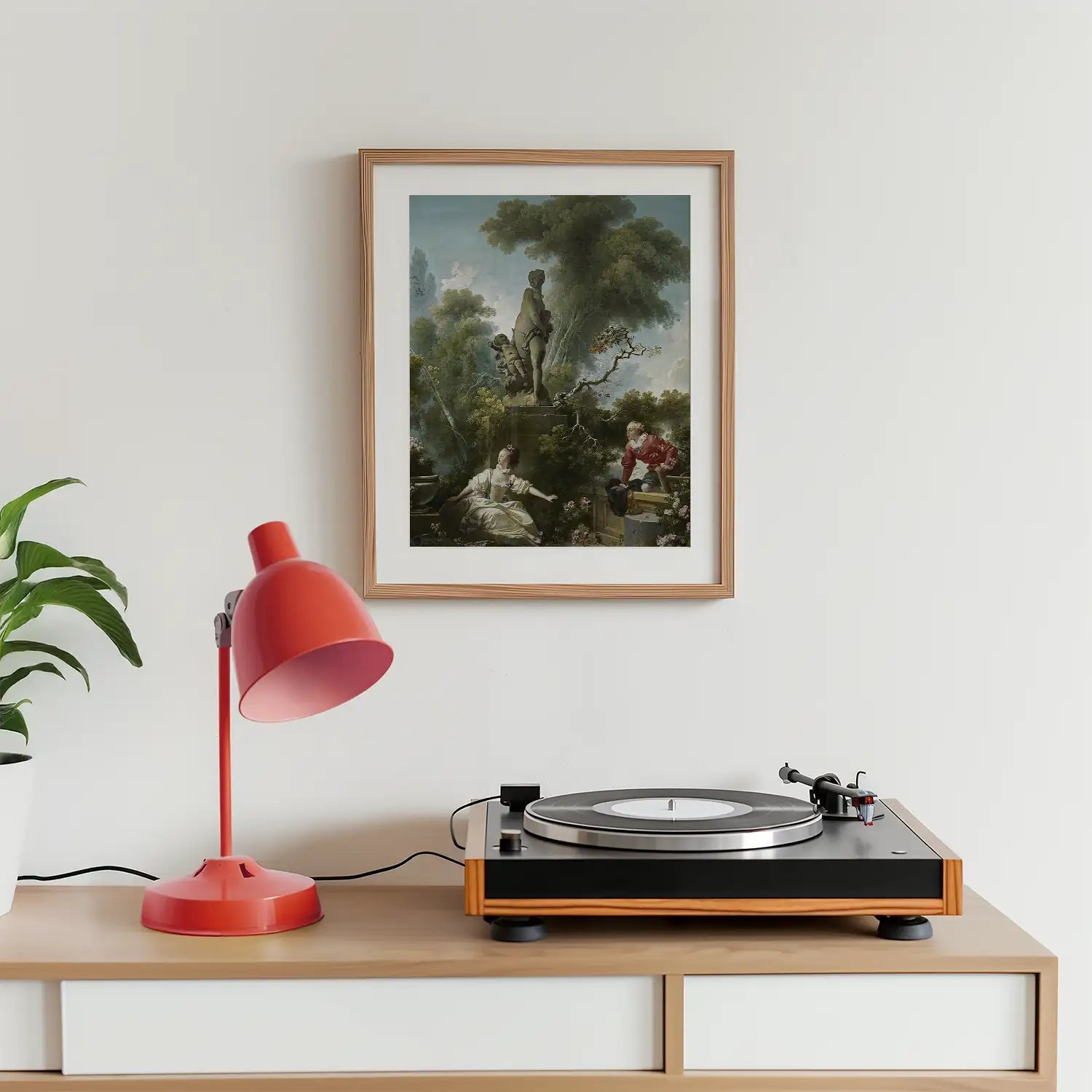 Framed painting of a serene outdoor scene with figures, displayed above a record player and a red desk lamp on a wooden surface.