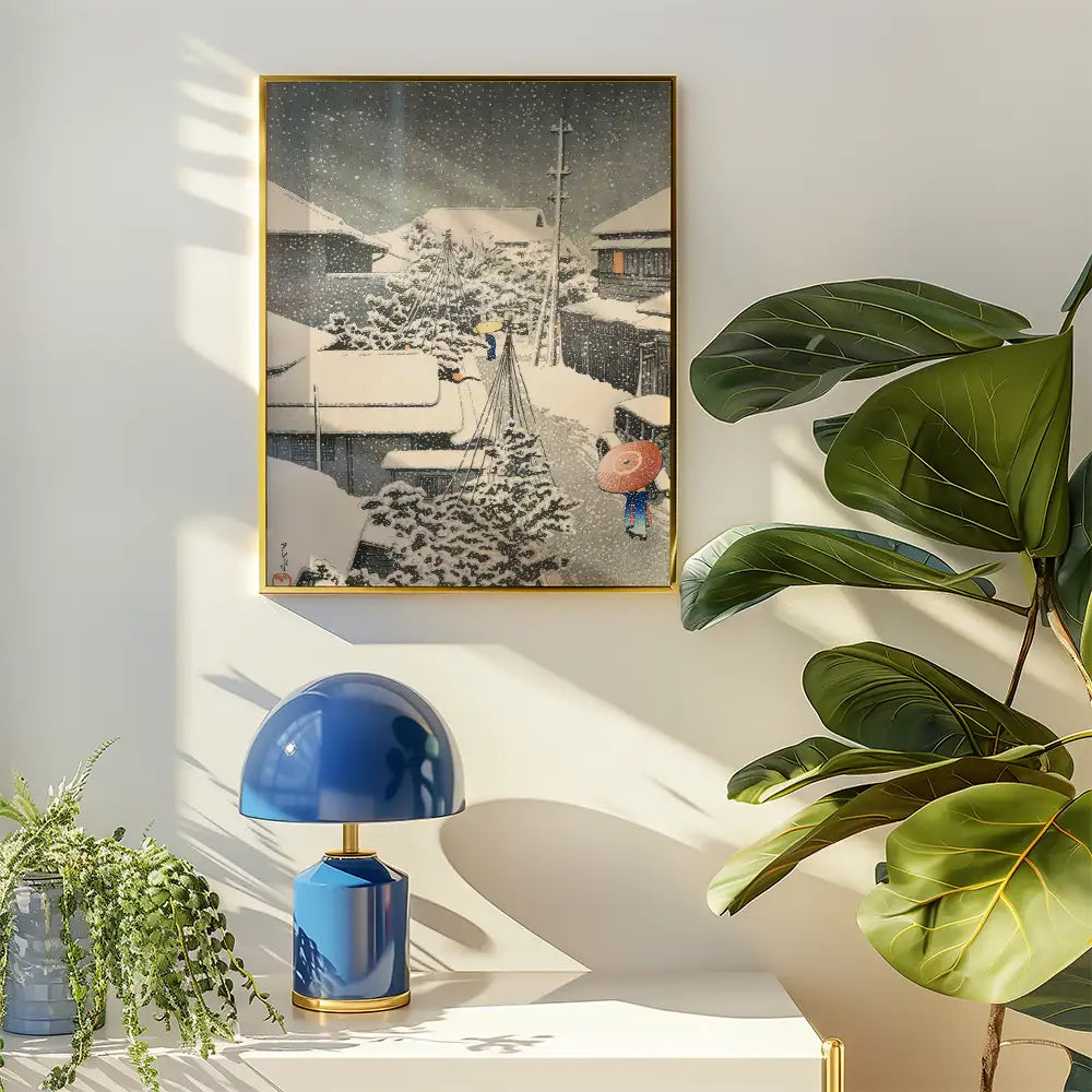 Framed painting of a snowy street scene with figures carrying umbrellas, displayed in a bright interior alongside a blue lamp and lush green plants.