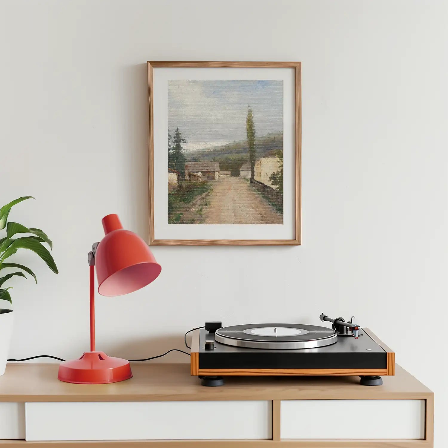A framed landscape painting hanging on a wall next to a red desk lamp and a record player on a wooden surface.