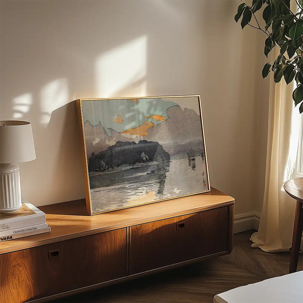 Framed landscape painting on a wooden surface with a lamp and plant in a bright, minimalistic room.