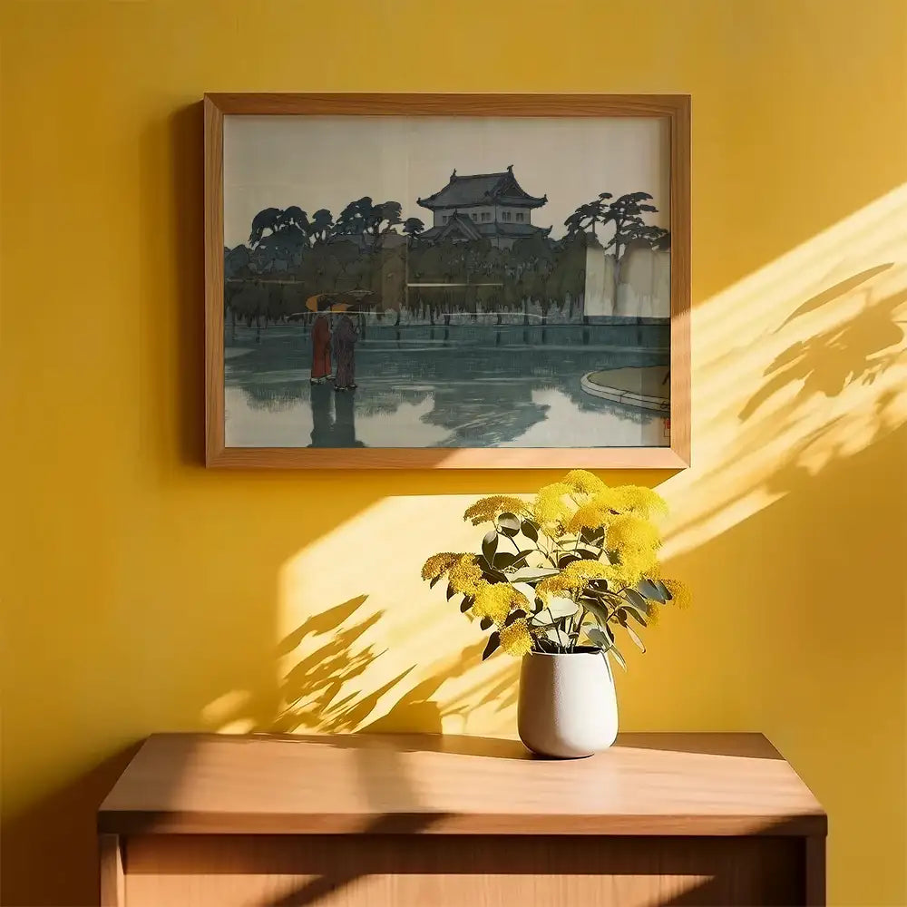 Framed painting of a serene landscape featuring two figures by a water body, displayed on a yellow wall with a potted yellow flower arrangement in front.