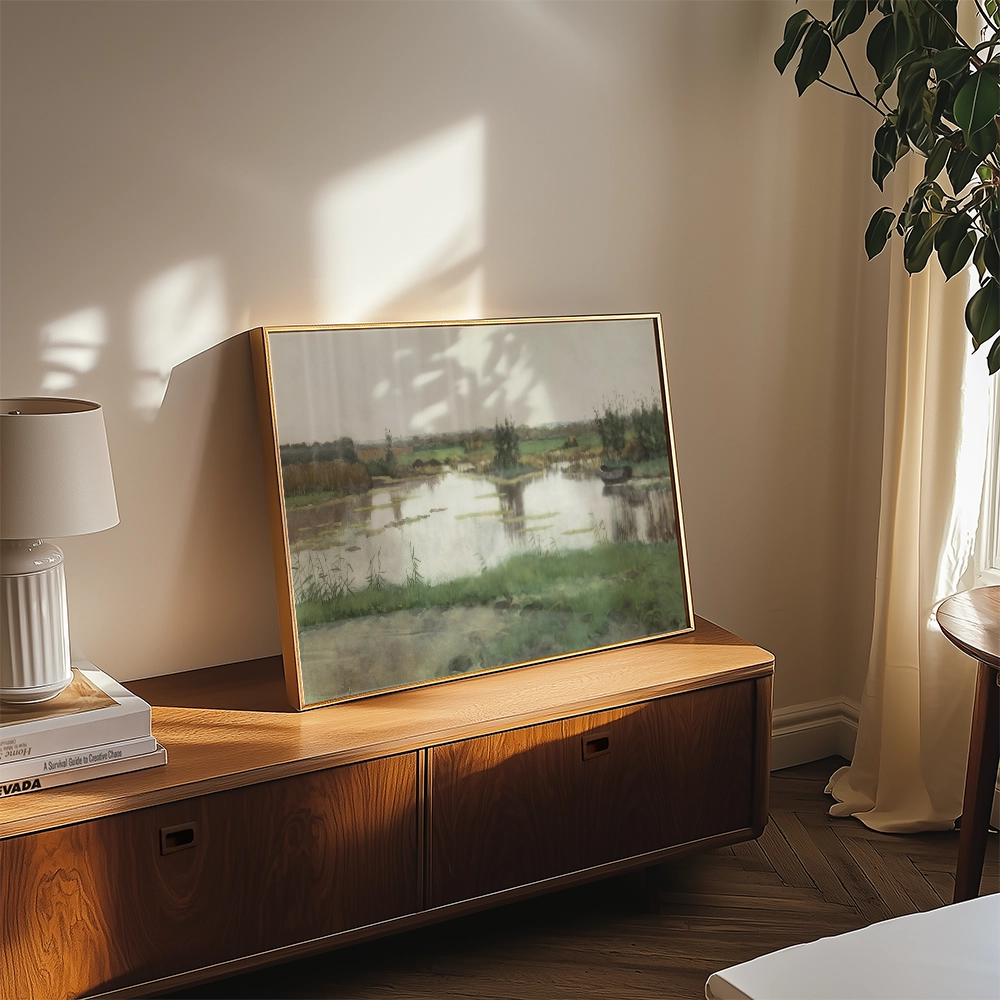 A framed landscape painting on a wooden side table with a lamp and decorative items in a sunlit room.