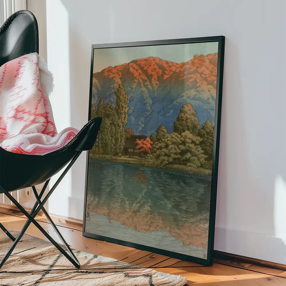 Framed painting of a serene landscape featuring mountains, trees, and a lake, placed next to a black chair and a patterned rug.