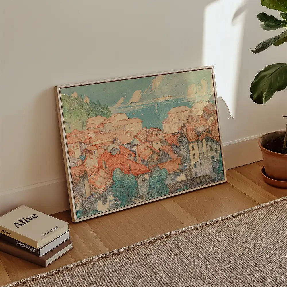 Framed painting of a coastal village with colorful rooftops placed against a neutral wall, alongside a potted plant and stacked books.