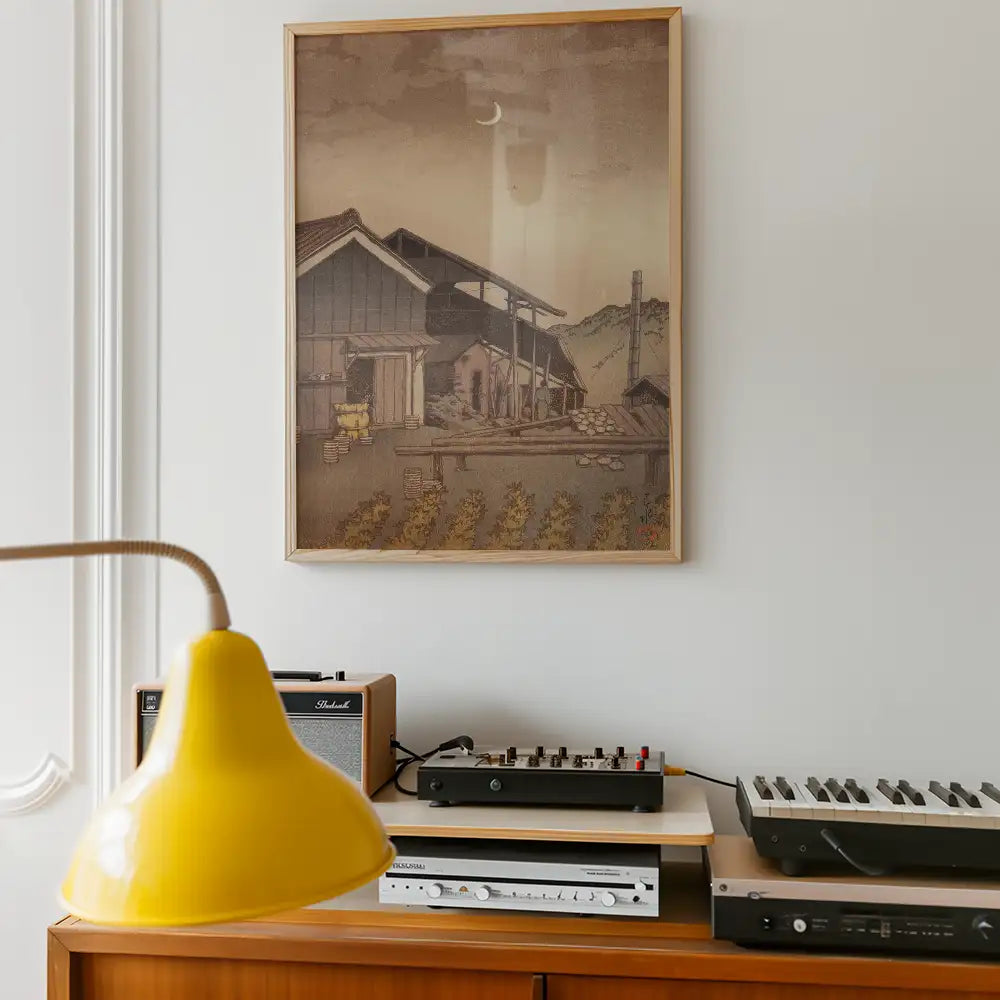 Framed painting of a rural landscape with houses and mountains, displayed on a white wall, alongside a yellow lamp and audio equipment.