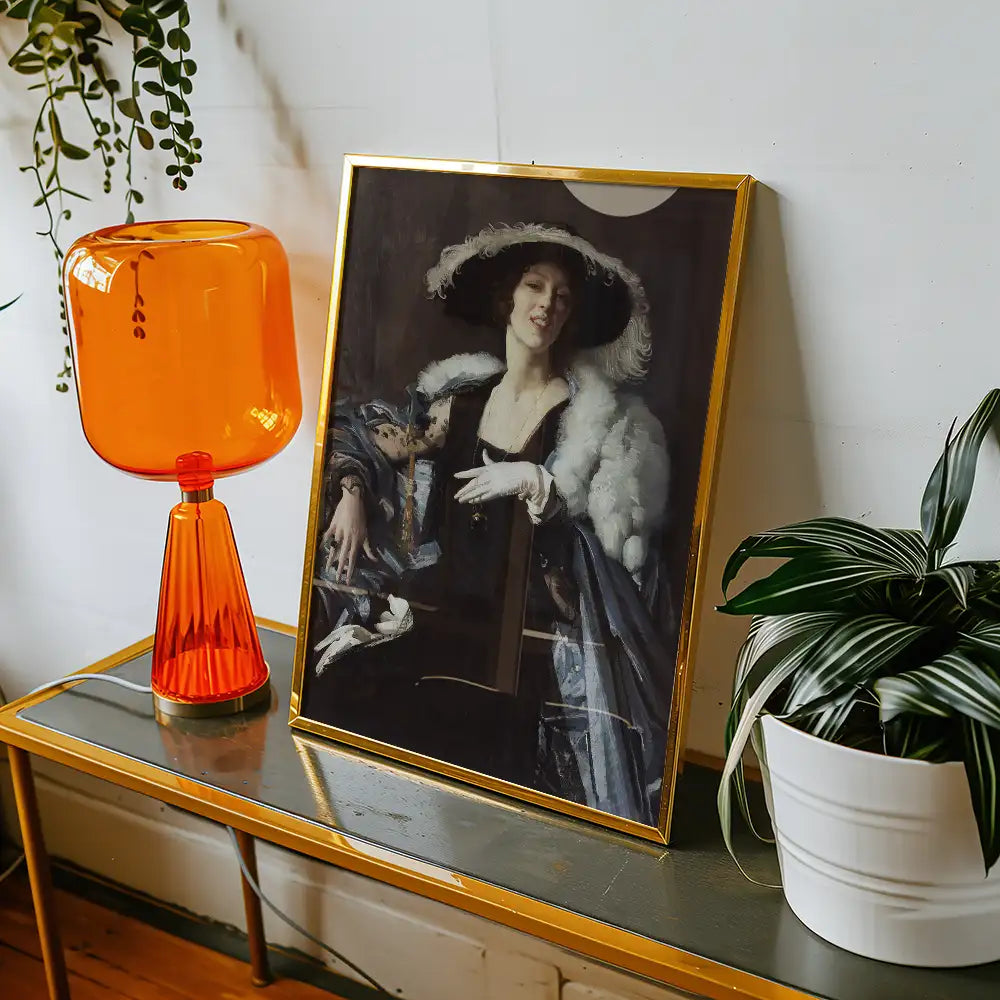 Framed painting of a woman in a large hat and fur shawl, displayed on a table next to an orange lamp and a decorative plant.