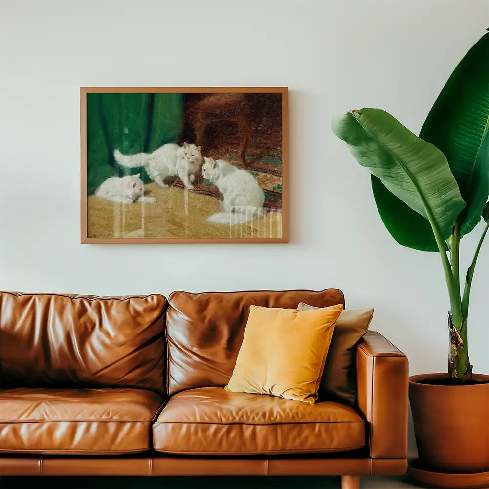 Painting of three white cats playing on a wooden floor, framed and hung on a wall above a brown leather couch with a yellow pillow and a green potted plant.