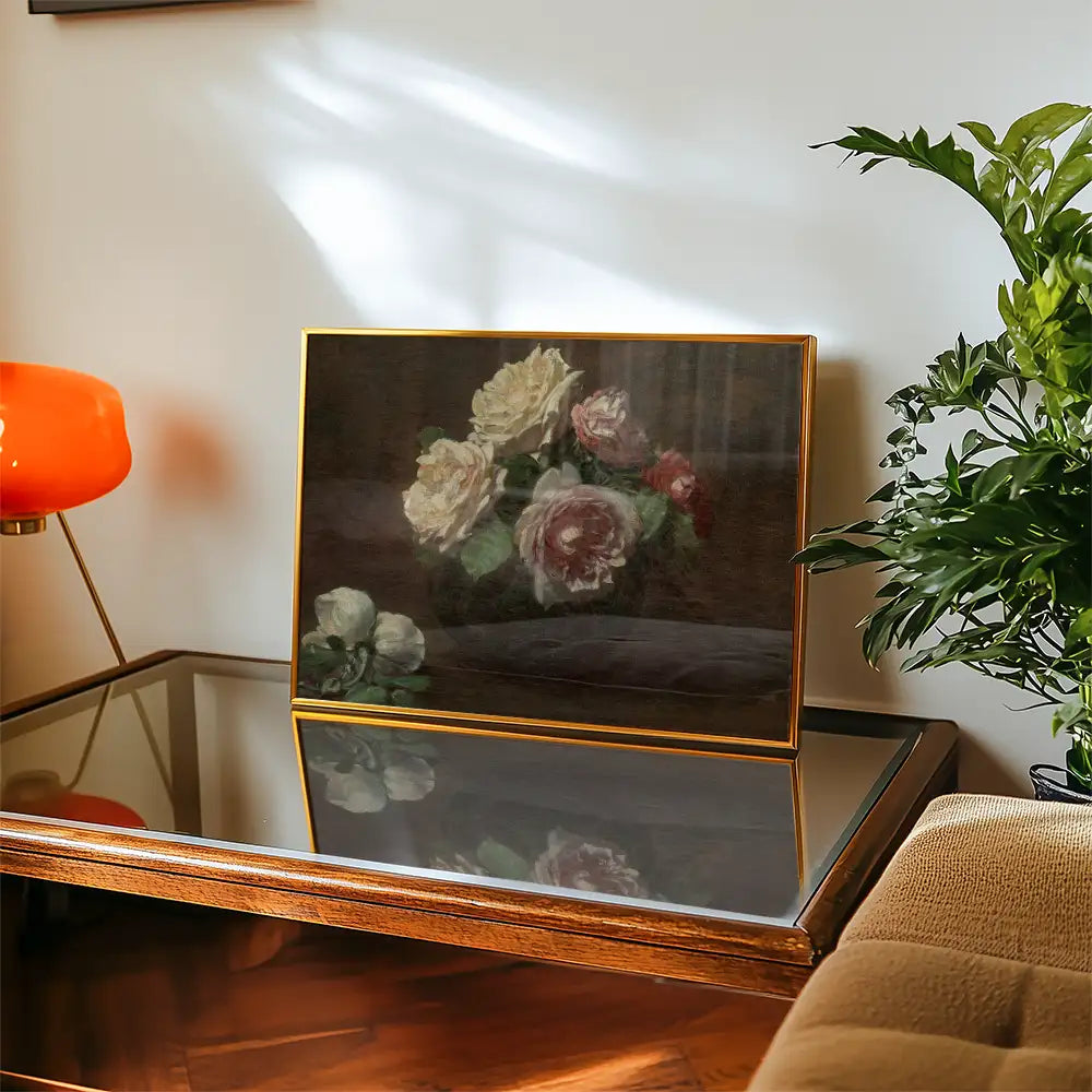 Painting of roses in a frame displayed on a wooden table with an orange lamp and a green plant in the background.