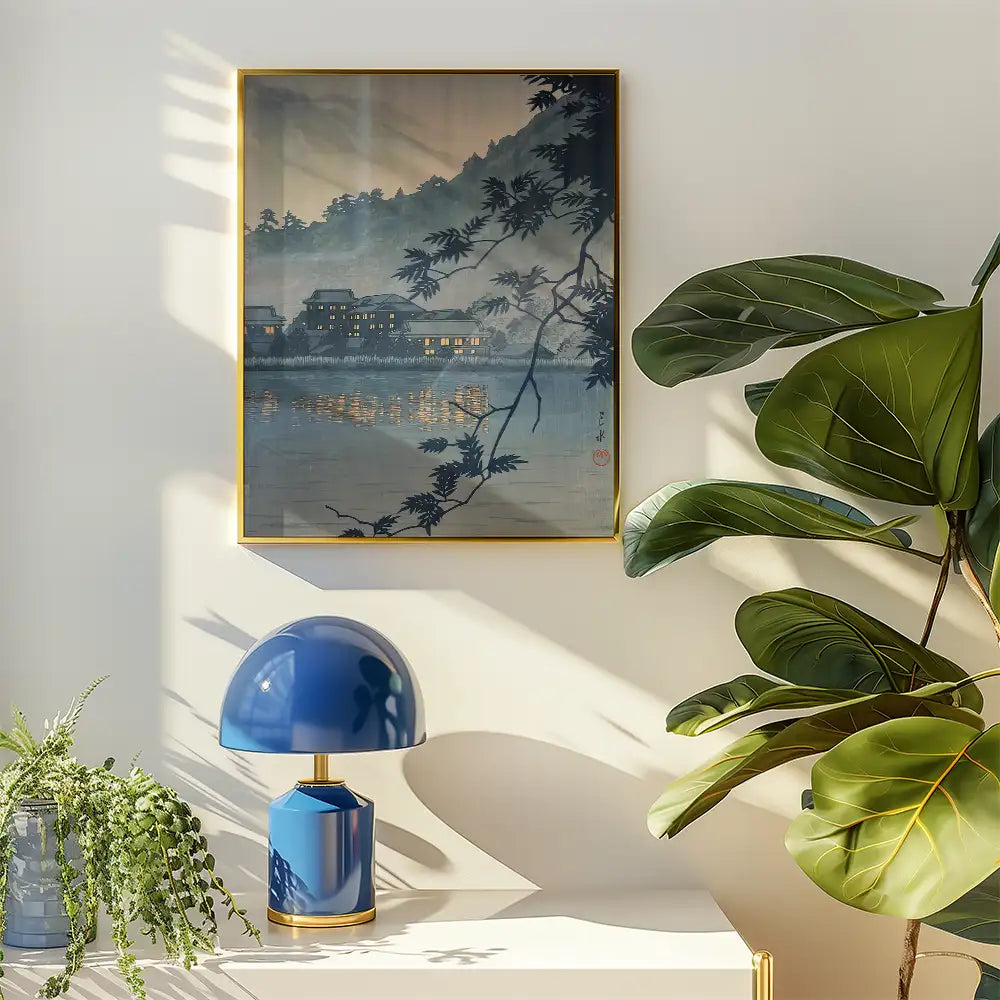 Framed painting depicting a serene lakeside scene with illuminated buildings, surrounded by dark foliage, next to a stylish blue lamp and green plant on a light-colored surface.