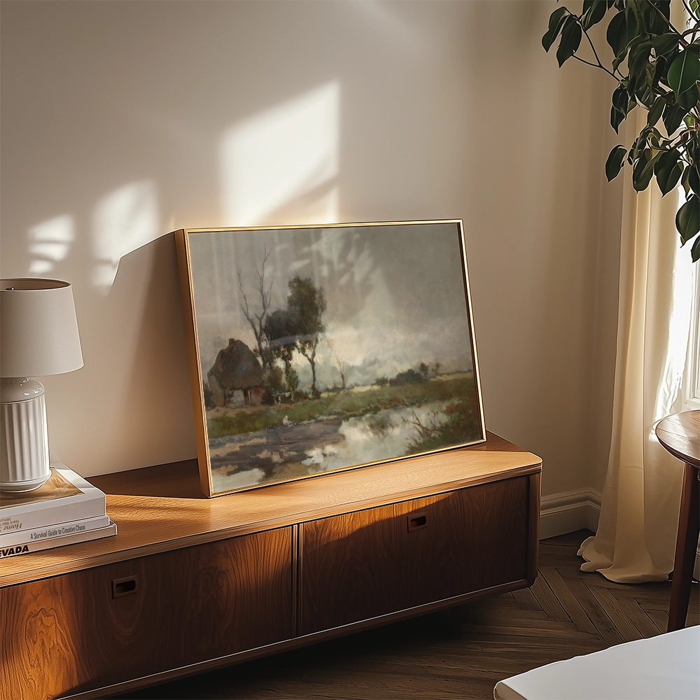A landscape painting in a golden frame displayed on a wooden console, with a lamp and books nearby, in a bright room with natural light.