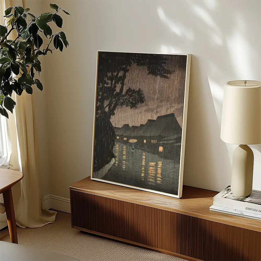 A framed painting of a rainy night scene with reflections on water, displayed in a cozy interior setting next to a lamp and plant.