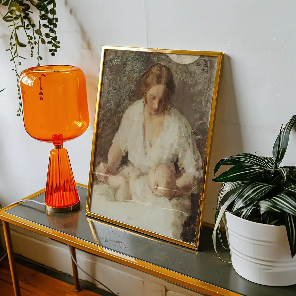 A framed painting of a woman holding two infants, displayed on a stylish table with a bright orange lamp and a potted plant.
