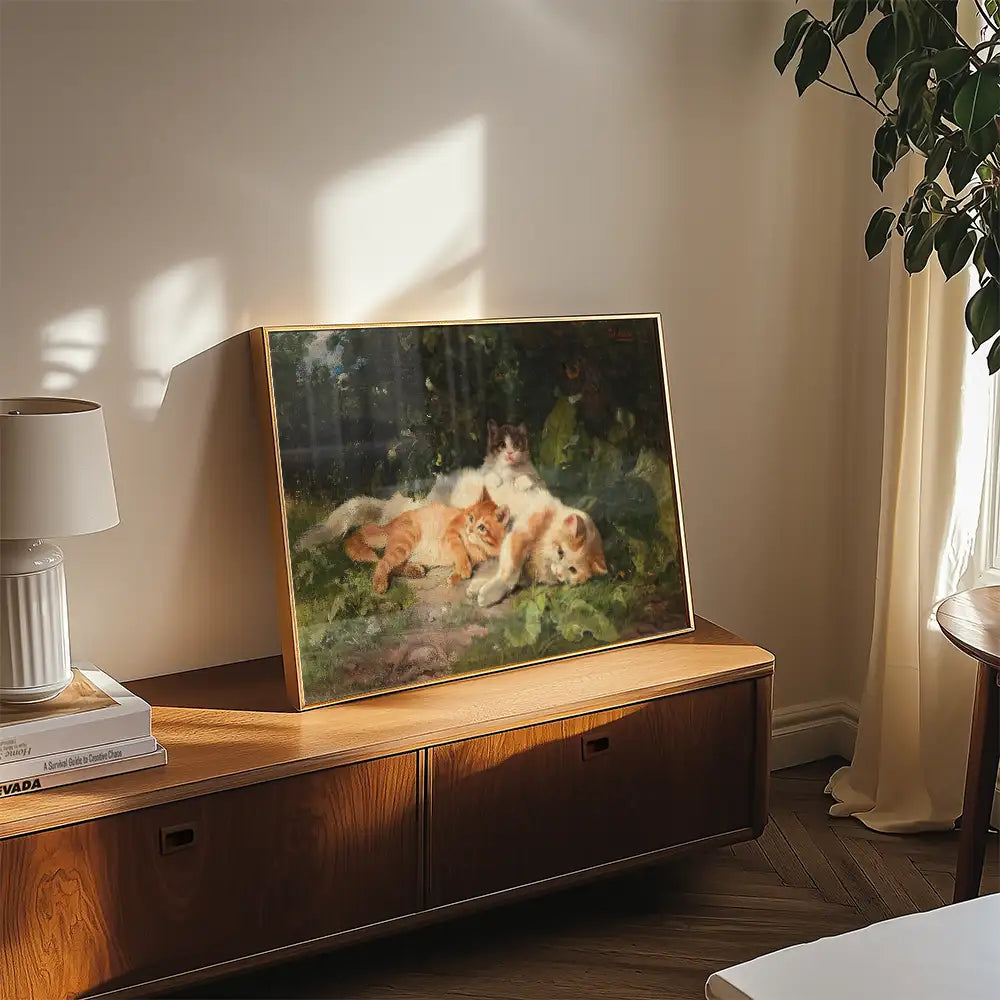 A framed painting of three cats resting on a grassy surface, featuring two orange cats and a white cat with black markings, displayed on a wooden table in a cozy interior setting with natural light.