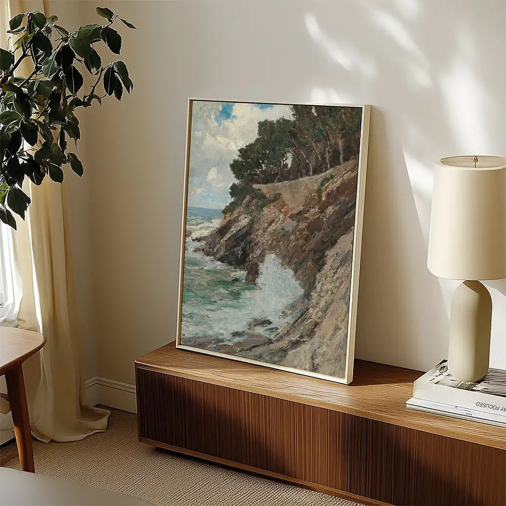 Framed painting of a coastal landscape displayed in a bright room with a plant and lamp in the background.