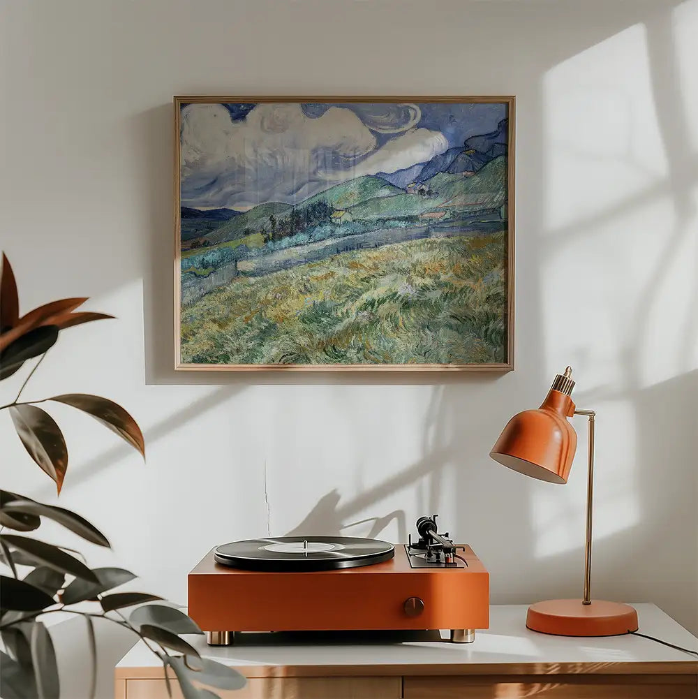 A framed painting of a landscape featuring rolling hills and cloudy skies, displayed above a modern orange record player and lamp in a cozy room setting.