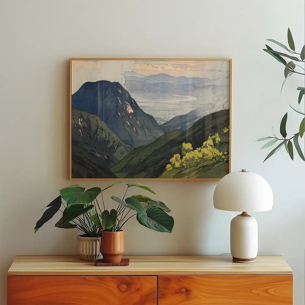 Framed landscape painting of mountains and green hills displayed on a wall above a wooden console table, accompanied by a potted plant and a decorative lamp.