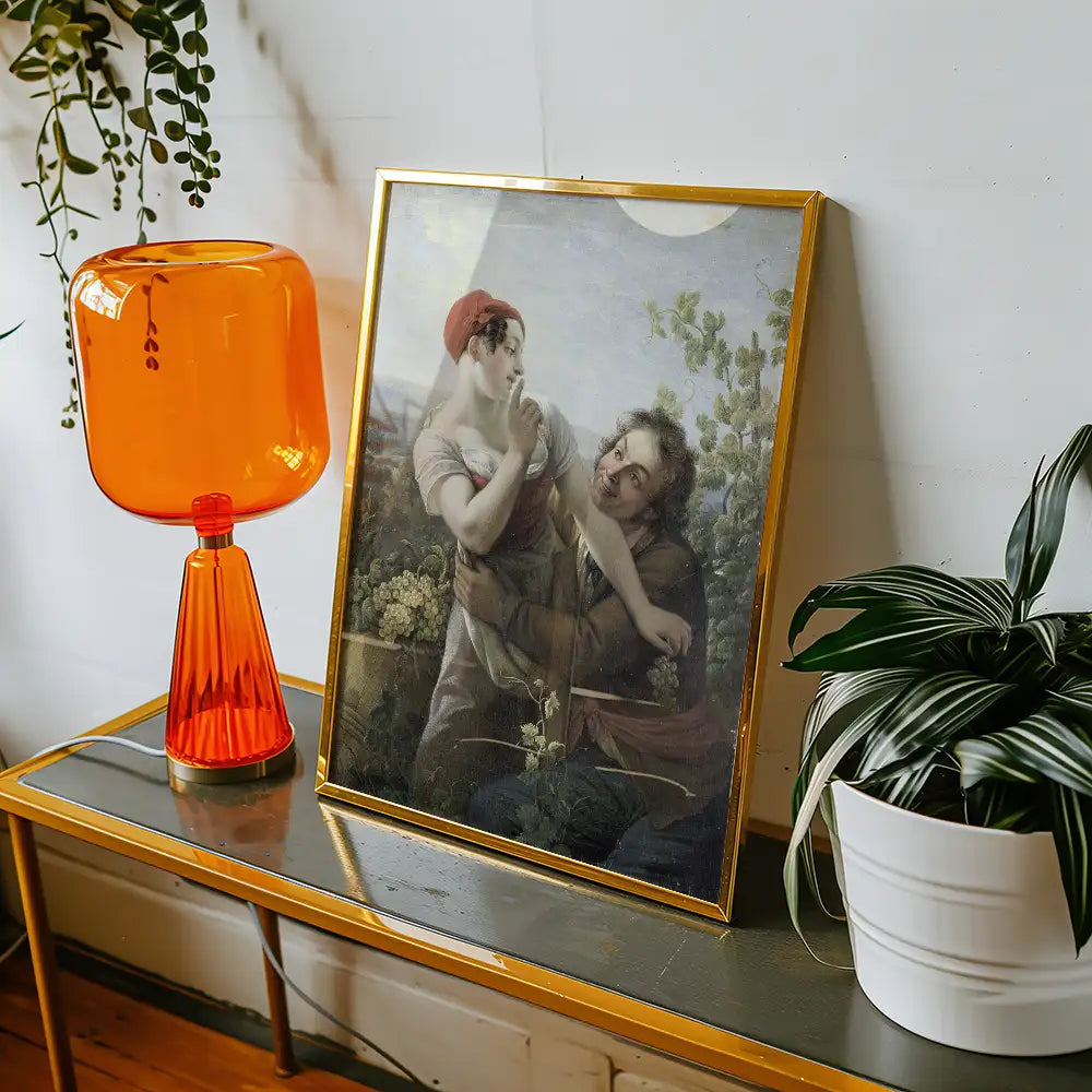 A vintage-style painting featuring a couple in a garden, framed in gold, displayed on a table beside an orange lamp and a potted plant.