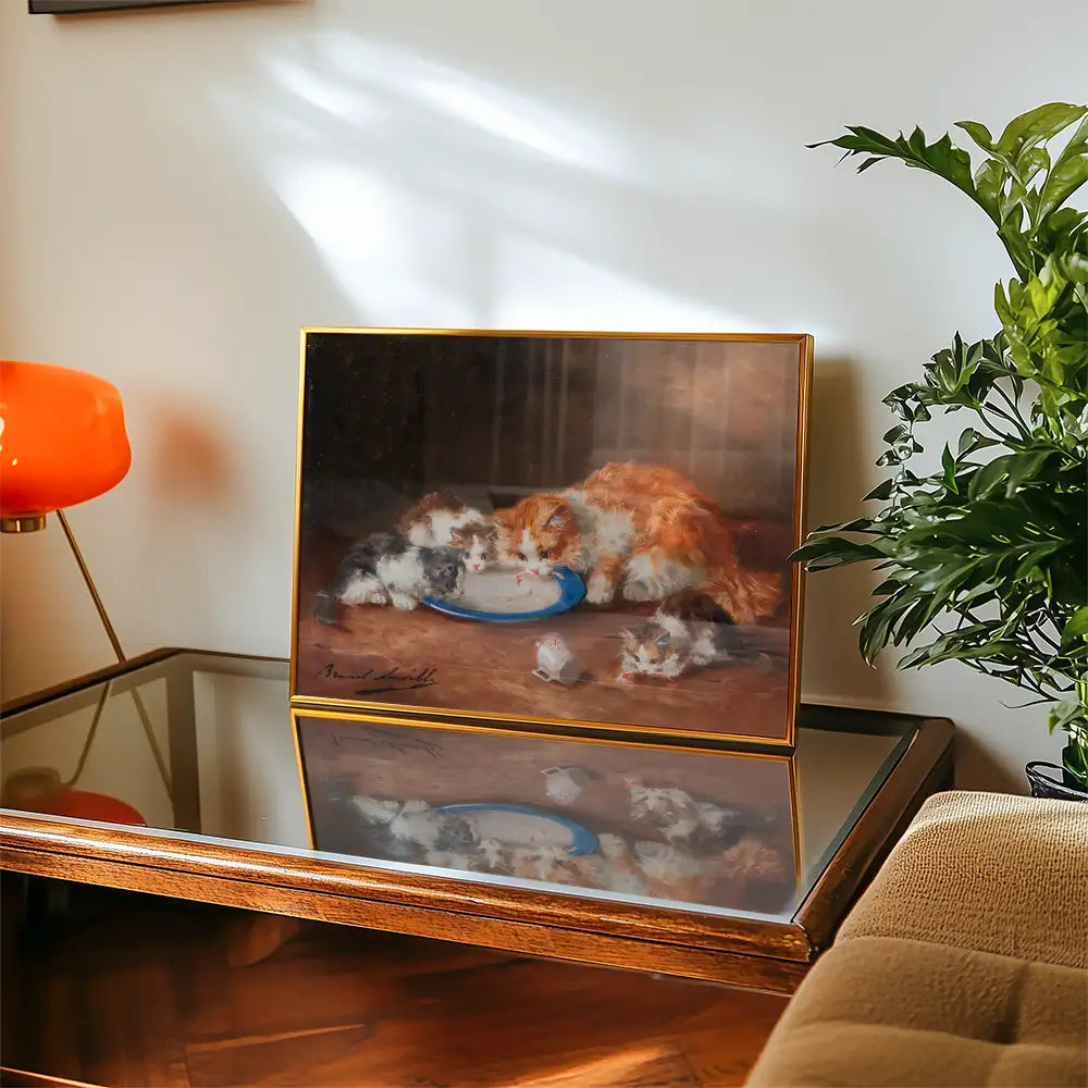 Painting of fluffy cats drinking from a blue plate, framed and displayed on a wooden table with a modern lamp and plant in the background.