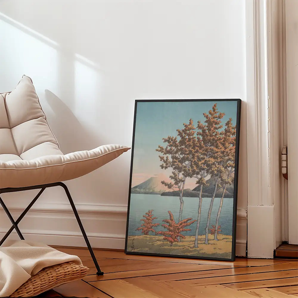 Framed painting of a serene landscape featuring trees by a lake with mountains in the background, leaning against a chair in a cozy room.