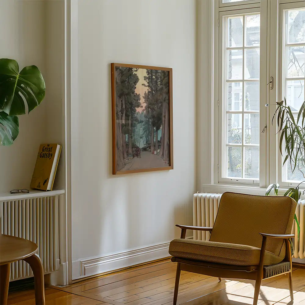 Framed painting of a forest scene hanging on a wall in a bright, modern living room with a chair and houseplant.
