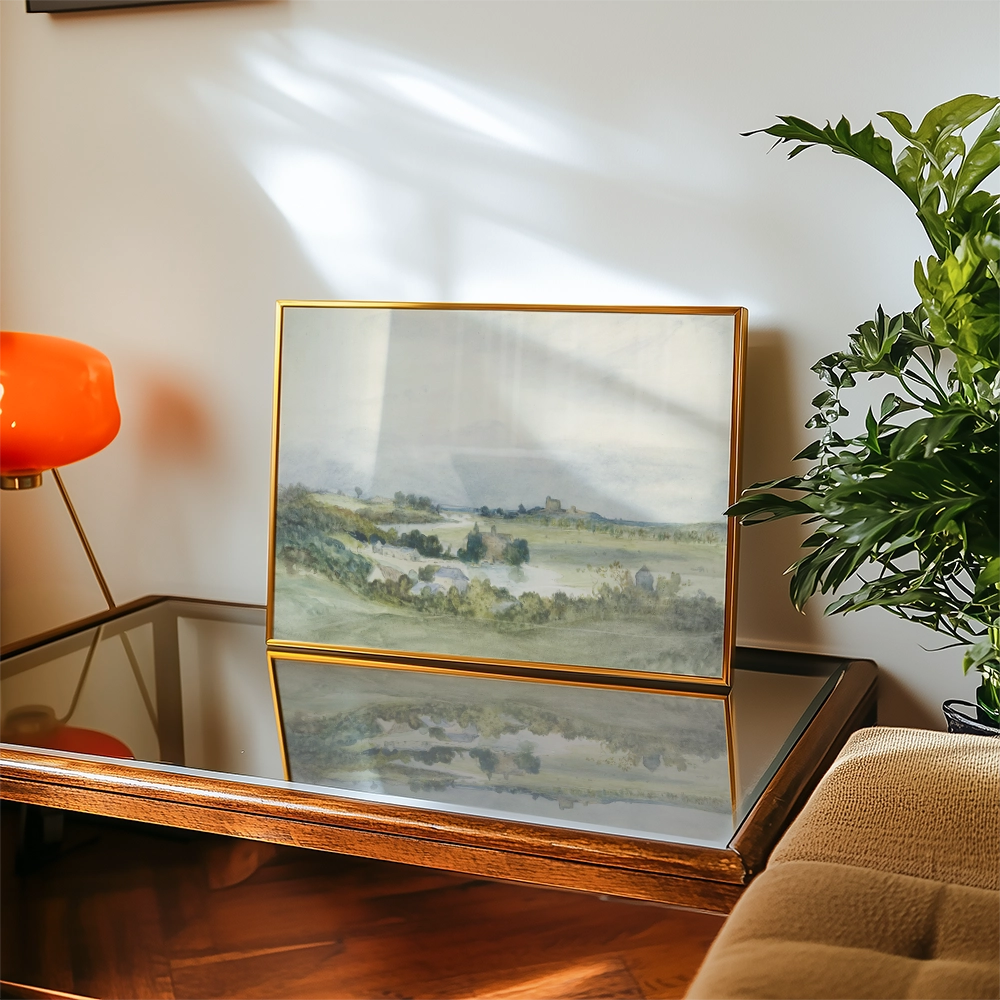 A framed watercolor landscape painting displaying rolling hills and a serene countryside scene, positioned on a glass-topped coffee table alongside an orange lamp and green foliage.