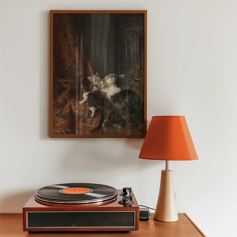 Painting of two cats playing on a rug, framed and displayed above a vintage record player and a modern orange lamp.