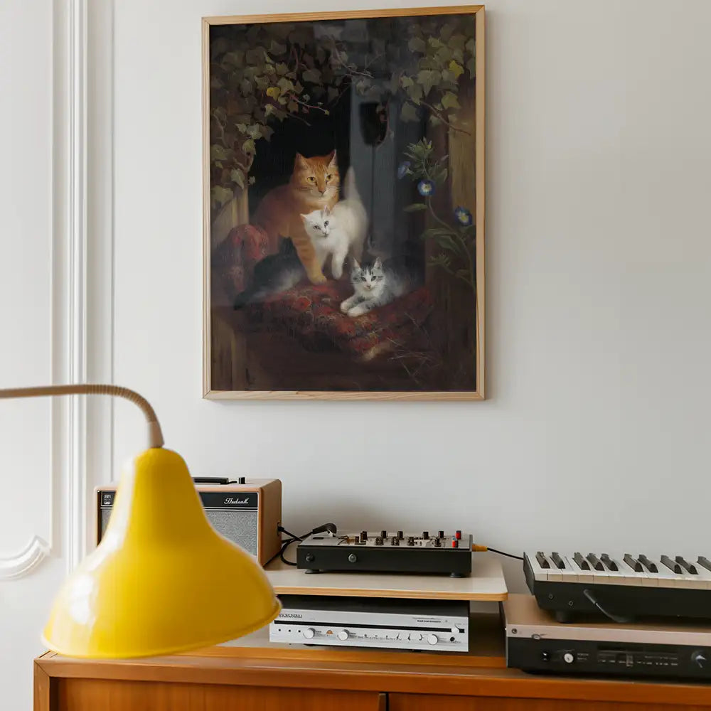 Painting of three cats in a cozy indoor setting, featuring an orange cat, a white cat, and a grey-and-white kitten, framed and hanging on a wall above a wooden shelf with electronic equipment and a yellow lamp.