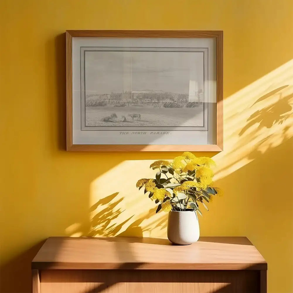 Framed vintage artwork titled "The North Parade" displayed on a yellow wall above a wooden cabinet with a vase of yellow flowers.