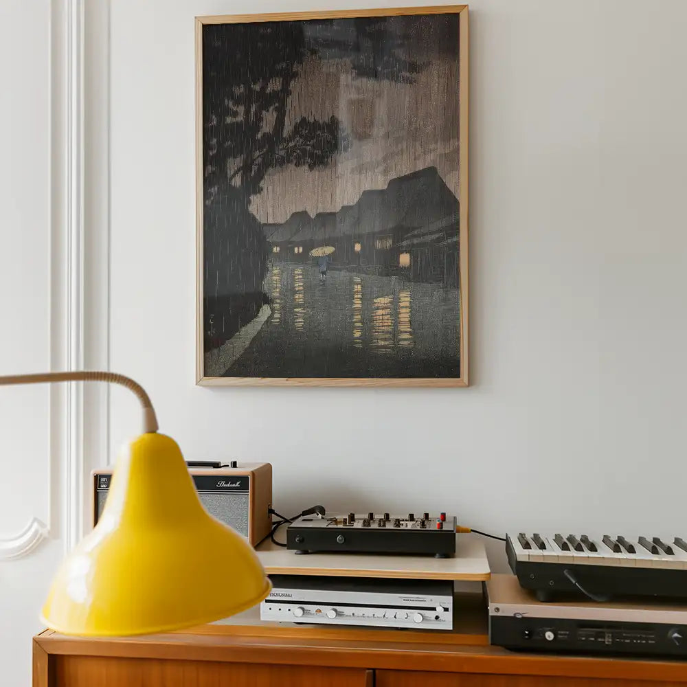 Framed painting of a rainy street scene with a person holding an umbrella, illuminated houses along the pathway, displayed on a wall above a wooden shelf with a yellow lamp and audio equipment.
