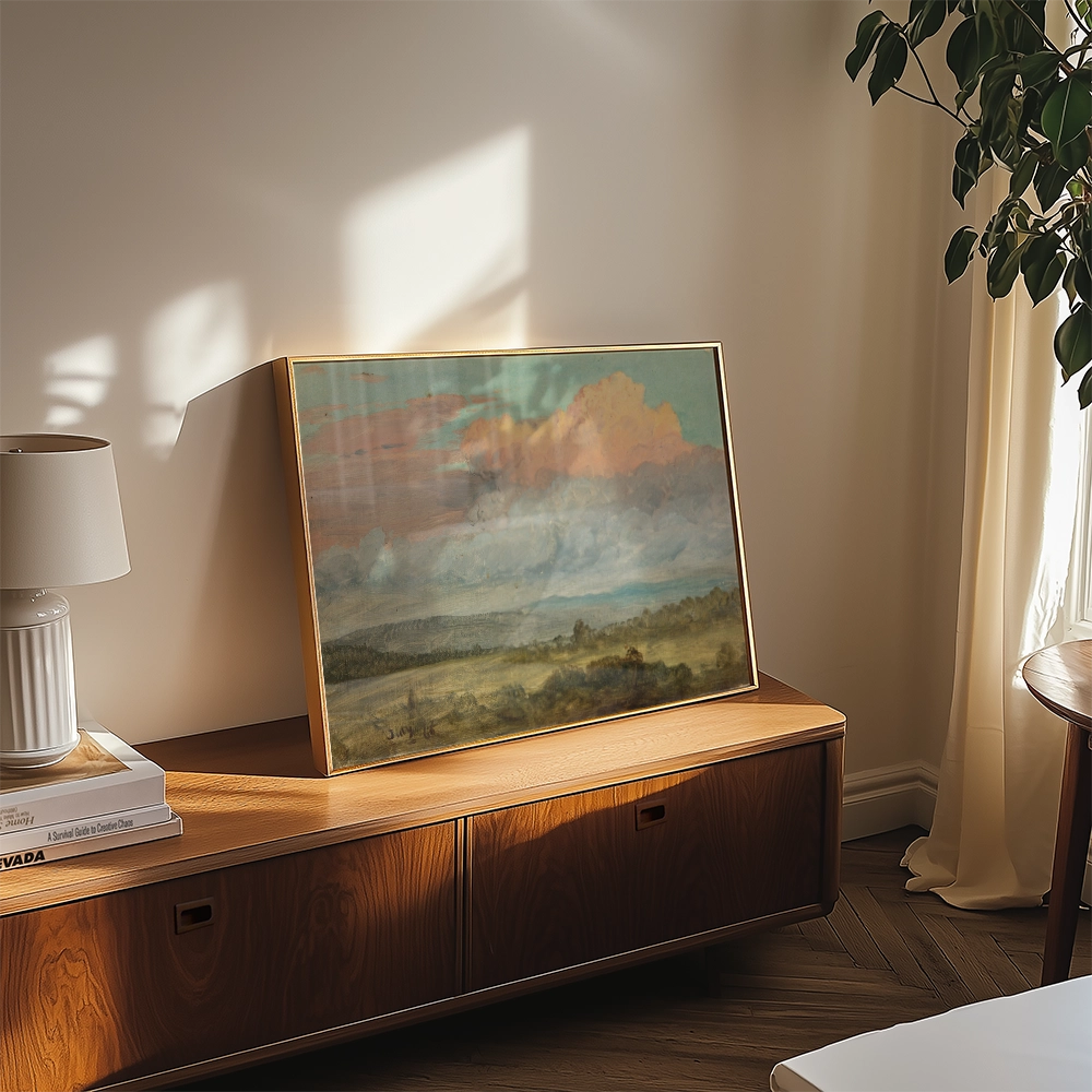 Framed landscape painting with soft clouds and hills displayed on a wooden console table beside a lamp and plant in a sunlit room.