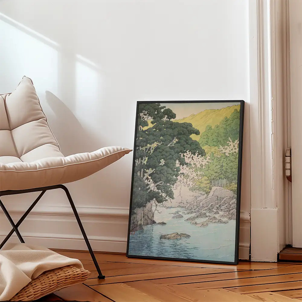 Framed painting of a serene landscape with a river, surrounded by trees and mountains, leaning against a wall next to a modern chair.