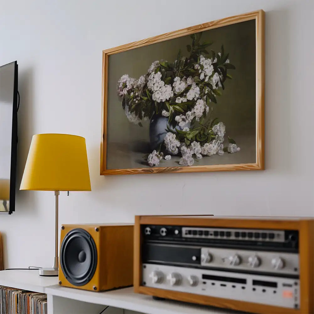 Framed painting of a bouquet of white flowers in a blue vase, displayed on a wall above a yellow lamp and a vintage audio system.