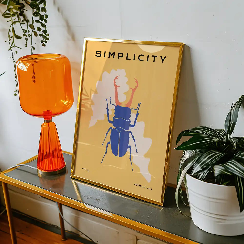 Modern art poster featuring a stylized blue beetle with the title "Simplicity," displayed in a gold frame next to an orange lamp and a potted plant.