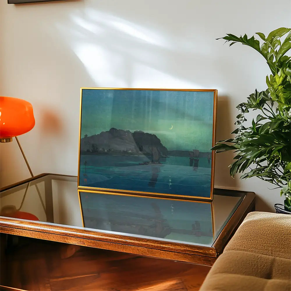 Framed painting of a serene landscape with water and mountainous terrain, displayed on a glass-topped table with a decorative plant and orange lamp in a cozy interior setting.