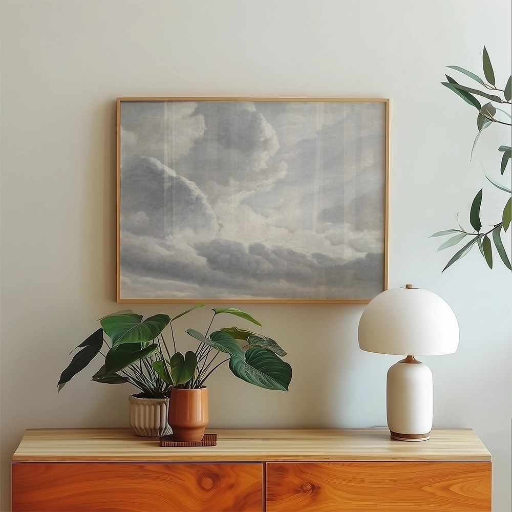 A serene painting of clouds in a light wooden frame, displayed above a wooden sideboard adorned with a potted plant and a modern lamp.