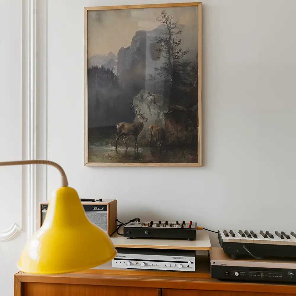 Framed painting featuring two deer near a rocky lake, surrounded by mountains and trees, displayed in a modern living space with musical equipment and a yellow lamp.