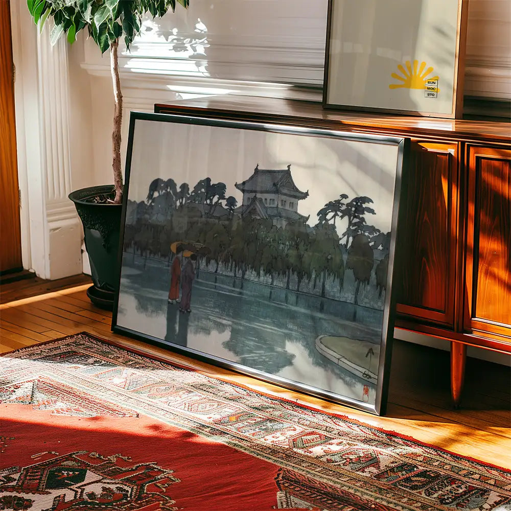 Framed painting of a serene landscape with figures holding umbrellas near a body of water, placed on a wooden floor beside a potted plant and a wooden cabinet.