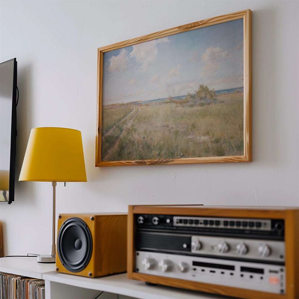 A landscape painting in a wooden frame depicting a grassy field under a blue sky with clouds, displayed on a white wall next to a yellow lamp and vintage audio equipment.