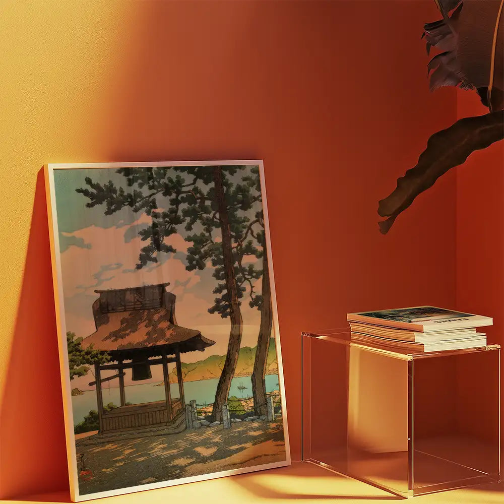 Framed painting of a serene landscape featuring a pagoda, trees, and a body of water, displayed against an orange wall with a stack of books on a clear side table.