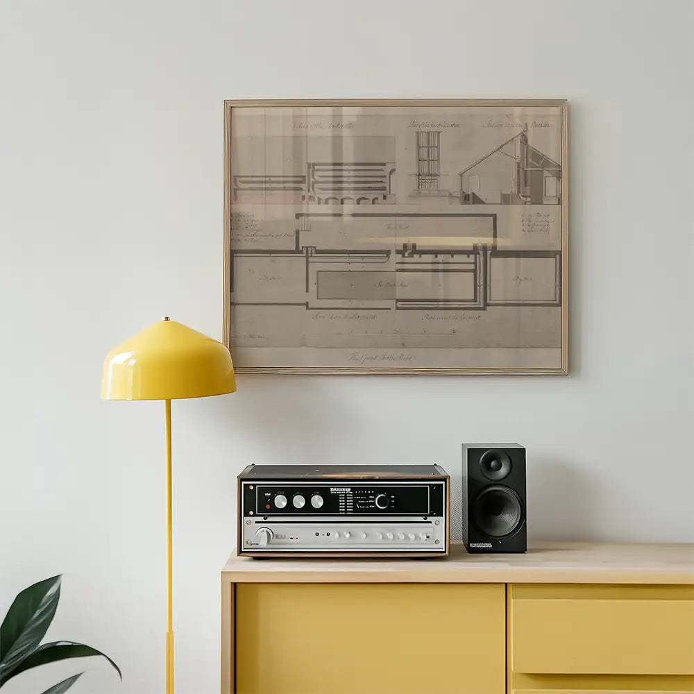 Vintage architectural blueprint framed art print hanging on a white wall above a yellow lamp and a sound system on a wooden console.