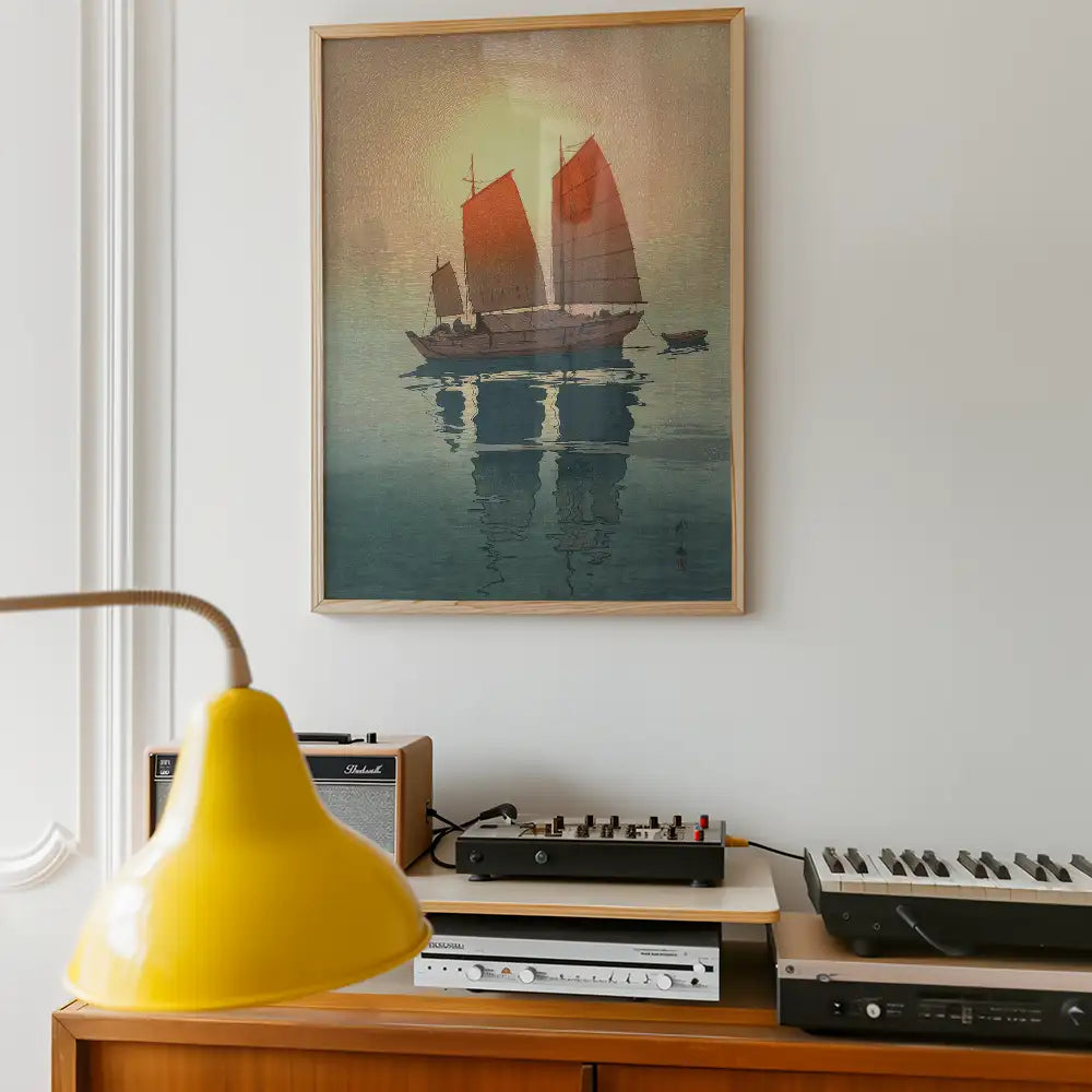 Painting of a traditional sailing ship with red sails reflected in calm waters, framed on a wall above a wooden console with music equipment and a yellow lamp.