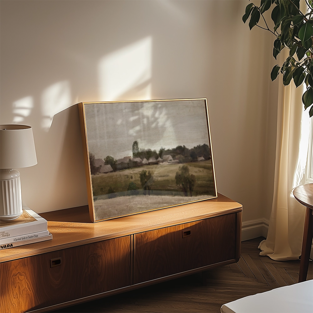 Framed landscape painting featuring softly blended colors, displayed on a wooden console table with a lamp and decorative books, illuminated by natural light.