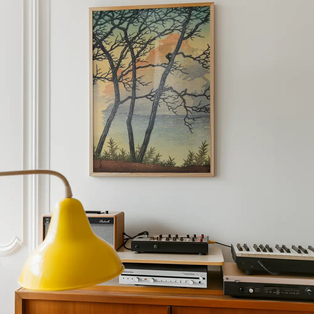Framed landscape painting of trees against a pastel background, displayed on a wall above a wooden side table with a yellow lamp and vintage sound equipment.