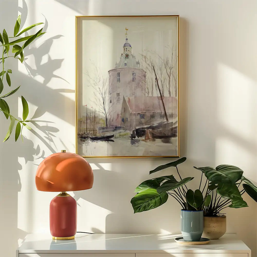 A soft watercolor painting of a tower and buildings by a body of water, framed in gold, displayed on a wall with a stylish orange lamp and indoor plants in a bright, sunlit room.