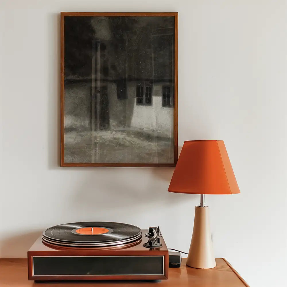 Painting of a dark, moody landscape with a house, displayed above a vintage record player and an orange lamp on a wooden table.