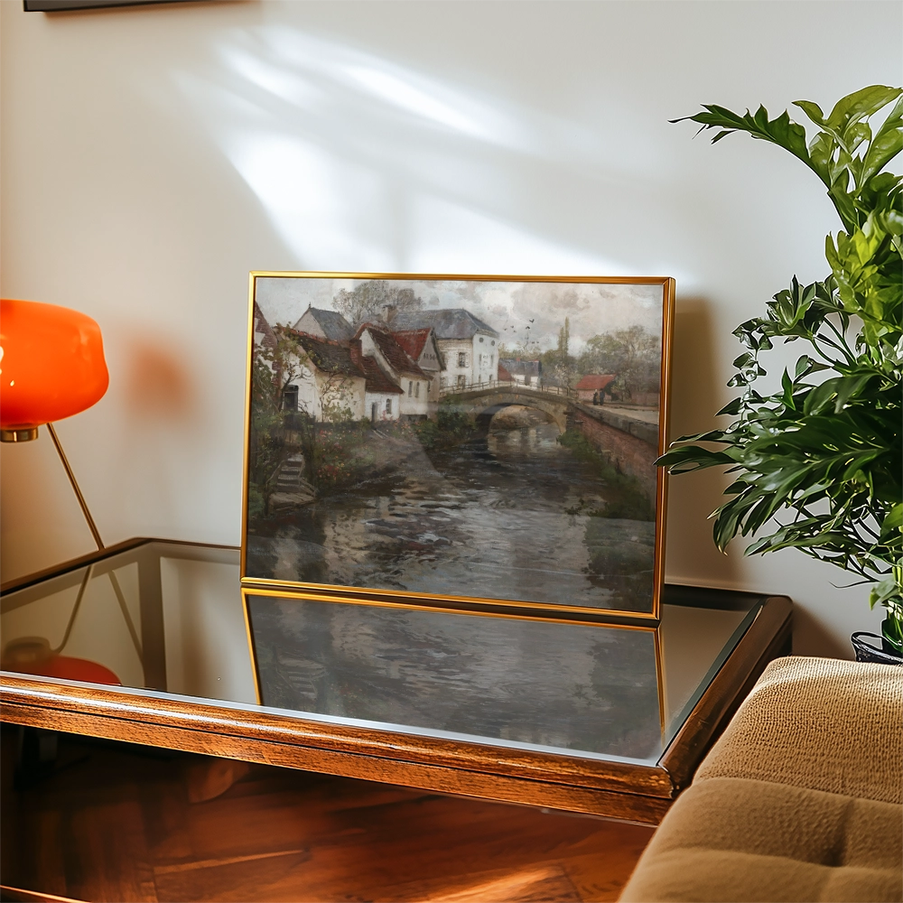 A framed painting of a serene river scene depicting houses and a bridge, displayed on a wooden table next to a potted plant and a modern lamp.